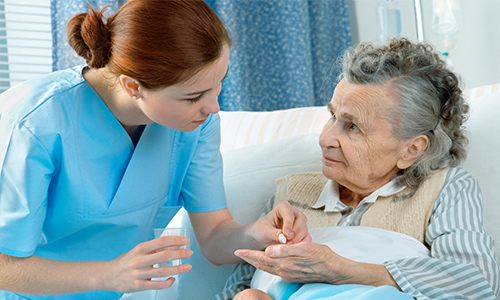 A caregiver sitting beside an older woman who is resting, talking with her and holding her hand.