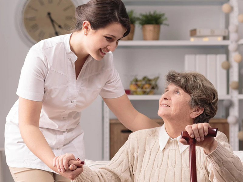 A caregiver helping an older woman adjust her cardigan as they sit together at home.