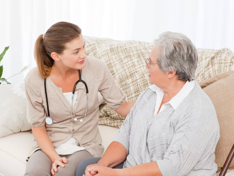 A caregiver sitting beside an older woman on a couch in a bright living room, talking with her.